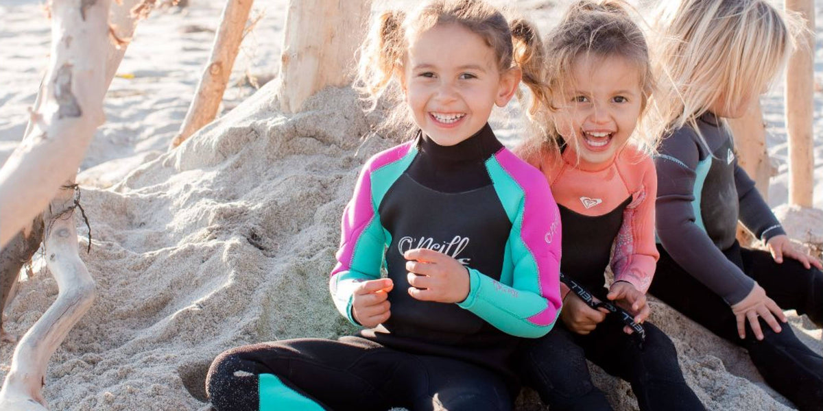 Neopren za otroke – deklice v dolgih otroških neoprenih sedijo na peščeni plaži